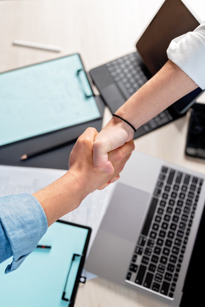 service-05 Two people shaking hands over laptops and documents in an office, symbolizing a business deal.